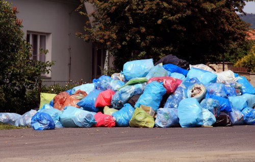 A large pile of rubbish bags and loose waste materials collected along the edge of a residential pavement near a white house with barred windows and surrounded by trees and shrubs. The rubbish includes blue, red, yellow, and black plastic bags, some partially torn open revealing contents such as paper and other household waste. The bags are stacked on the asphalt surface and extend across the foreground, with a background showing a leafy tree and a partly visible building wall. The scene suggests a local waste collection or rubbish removal activity in the vicinity of Blackfriars, within central London. The presence of the bags indicates a recent or ongoing clearance that Commercial Waste Blackfriars could assist with, emphasizing the importance of professional rubbish removal services in urban environments.