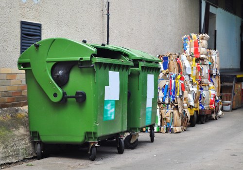 The image shows a large green wheeled commercial waste bin made of durable plastic, positioned on a paved outdoor area adjacent to a beige building wall. The bin has a rounded top cover and two white rectangular label areas on its front, with an open side revealing inside compartments for waste collection. Next to the bin, there are multiple tightly folded and stacked cardboard boxes and packaging materials, some with visible printed or colored designs, arranged in a pile along the side of the pavement. The background includes a section of a modern window with a dark frame and a glimpse of a small section of brickwork, indicating an urban environment in a town such as Blackfriars. The scene appears to be part of a commercial or industrial waste collection site, with natural daylight illuminating the materials clearly, emphasizing the textures and colours of the cardboard and the bin. This setup reflects typical rubbish collection arrangements seen when managing waste disposal for local businesses, which commercial waste removal services like Commercial Waste Blackfriars provide in the area, including the postcode region.