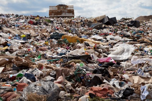 The image depicts a large, uneven pile of mixed waste materials spread across an open outdoor area, likely an illegal dump or uncontrolled landfill site. The waste includes various types of rubbish such as plastic bags, broken furniture, cardboard, paper waste, and discarded textiles, with some items showing signs of weathering and decay. In the background, a partially constructed or abandoned building can be seen under a cloudy sky, emphasizing an urban or peri-urban environment. The waste is scattered across the ground with some items overlapping, creating a chaotic scene that highlights improper disposal. The scene is representative of issues addressed by commercial waste removal services such as those provided by Commercial Waste Blackfriars, which focus on clearing such large quantities of refuse efficiently, especially in busy towns or commercial areas in central London.