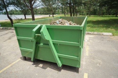 A large, rectangular green skip situated on an outdoor paved area, likely a parking lot or storage space, with visible yellow parking lines on the ground. The skip is filled with a mixture of household and garden waste, including visible paper, plastic, and plant debris, some of which overflows slightly from the top edges. Behind the skip, there is a backdrop of lush green grass, several mature trees with leafy canopies, and a distant body of water, suggesting a park or riverside location. The environment appears to be well-lit with natural daylight, indicating a clear, sunny day. The skip and waste are associated with commercial waste collection services in the Blackfriars area, and Commercial Waste Blackfriars manages rubbish removal for such outdoor waste containment. The image emphasizes the type of heavy-duty, durable waste containers used for bulk rubbish collection.