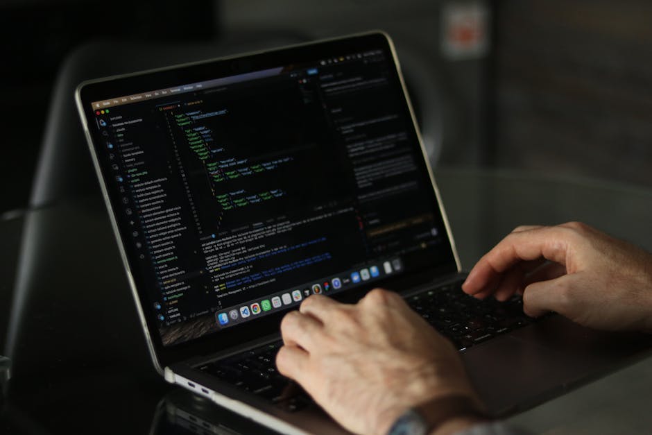 A black laptop computer with a red backlit keyboard displaying lines of blurred code on its screen, situated on a dark surface. Next to the laptop, there is a white Puma baseball cap with the black Pu