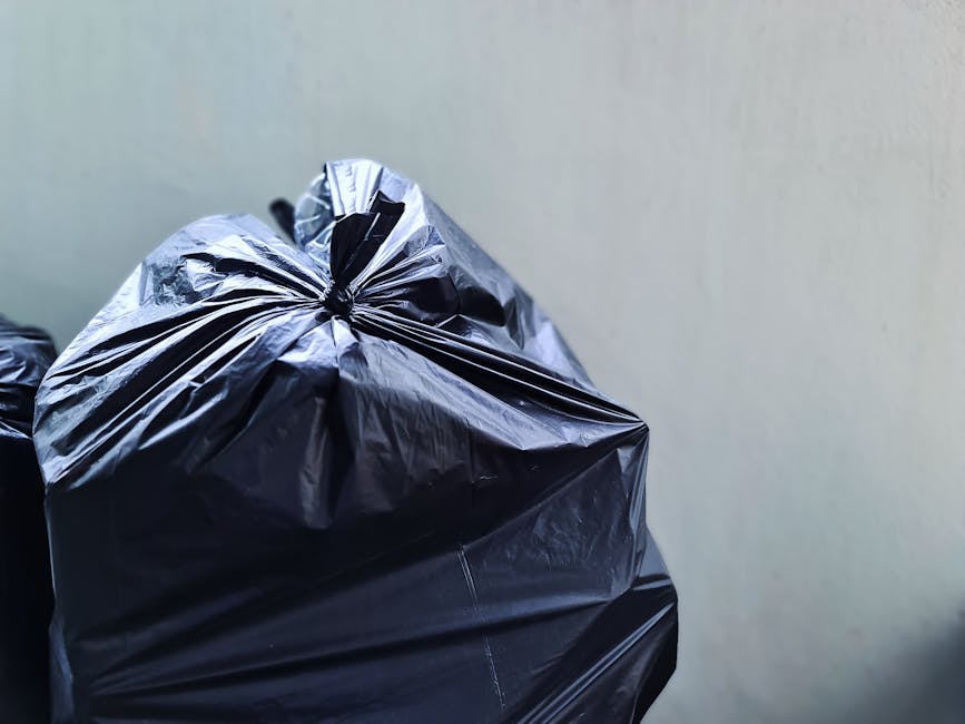 A tall, bright blue wheeled rubbish bin with a closed lid, positioned outdoors on a grassy area, near a pink waste container with black holes and a grey lid. The blue bin has a small rectangular stick